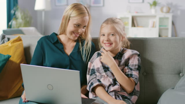 Schöne-junge-Mutter-und-ihre-niedlichen-kleinen-Tochter-Verwendung-Laptop-beim-Sitzen-auf-einem-Sofa-zu-Hause.-Familie-verbringen-Zeit-zusammen-betrachten-von-Videos-und-Cartoons-auf-Computer.