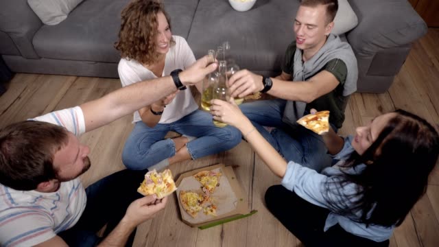 Friendship,-food,-domestic-party-time---cheerful-caucasian-friends-order-pizza,-having-fun,-sitting-on-the-wooden-floor-indoors-and-laughing,-eating,-drinking,-cheering.-Top-view