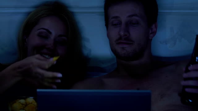 Young-couple-in-bed-watching-serials-on-laptop-eating-snacks-and-drinking-beer