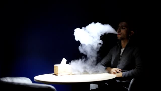 Young-man-sitting-on-the-bar-smoking-e-cigarette-vape-in-the-dark-cafe