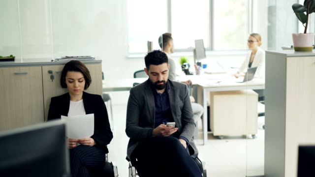 Young-people-man-and-woman-are-waiting-for-job-interview-in-office-while-manager-is-interviewing-another-candidate.-Girl-is-holding-cv,-guy-is-using-smartphone.