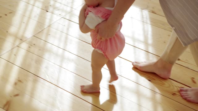Hohe-Winkelansicht-von-niedlichen-Mädchen-in-rosa-Bodysuit-macht-ihre-ersten-Schritte-durch-die-Pflege-Mama-zu-Hause-unterstützt