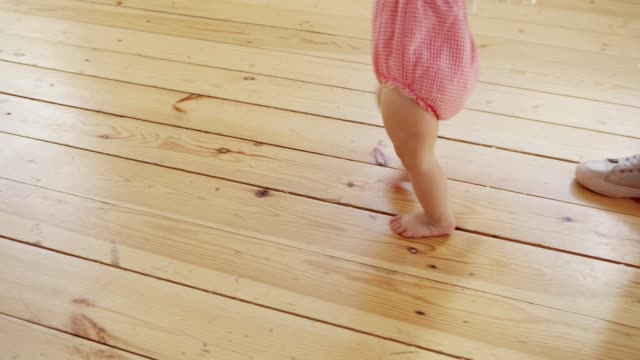 Tracking-shot-of-mother-supporting-her-adorable-baby-daughter-in-pink-bodysuit-learning-to-walk-on-hardwood-floor-at-home