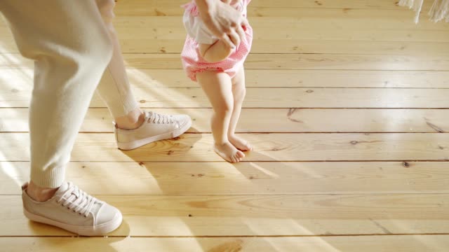 High-angle-view-of-cute-baby-girl-learning-to-walk-on-wooden-floor-at-home-helped-by-her-caring-mother-in-slow-motion