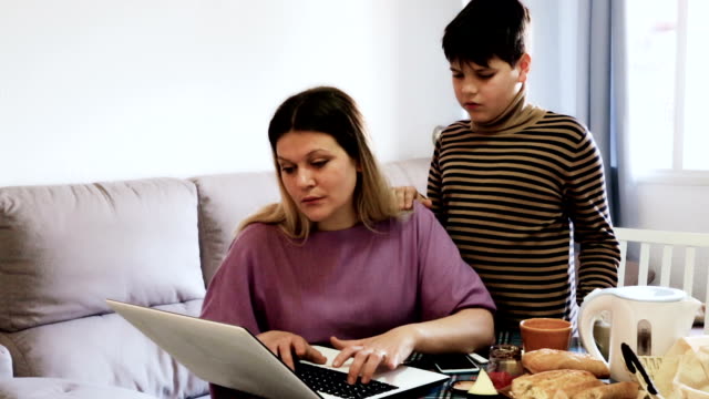 woman-working-at-laptop-and--sad-preteen-boy-standing-near-table