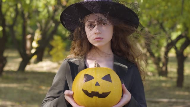 Ernsthaft-aussehende-Rothaarige-kaukasische-Mädchen-in-Halloween-Hexenkostüm-im-Herbstpark-stehend.