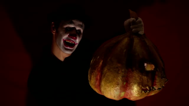 Scary-clown-holds-a-pumpkin-mask-in-his-hands.-A-scary-man-in-a-clown-makeup-holds-a-pumpkin-for-Halloween.-A-scary-clown-holds-a-Jack-O-Lantern-in-his-hands-with-luminous-eyes-and-a-mouth.