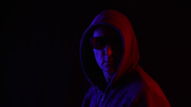 Portrait-adult-man-in-black-sunglasses-and-hoodie-turning-around-in-studio-on-black-background.-Man-in-hood-on-black-background-in-blue-and-red-lighting-looking-to-camera.
