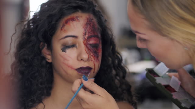 Make-up-artist-make-the-girl-halloween-make-upin-studio.Halloween-face-art.Woman-applies-on-professional-greasepaint-on-the-face-of-spanish-girl.War-paint-with-blood,-scars-and-wounds.Slow-motion