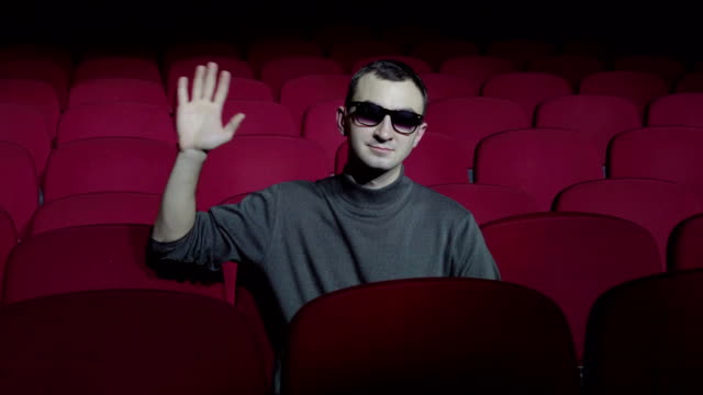 Single-man-sitting-in-comfortable-red-chairs-in-dark-cinema-theater-and-waving-hand-to-camera