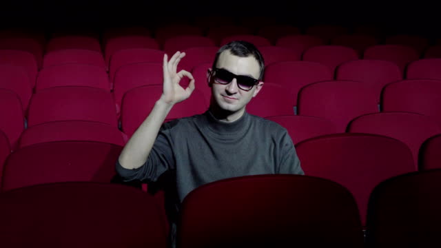 Single-man-sitting-in-comfortable-red-chairs-in-dark-cinema-theater-and-showing-ok-sign