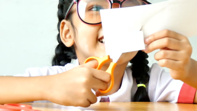Kleine-Asiatin-in-Thai-Kindergarten-Studenten-Uniform-mit-Schere-um-zu-schneiden-Sie-das-White-Paper-auf-Holztisch