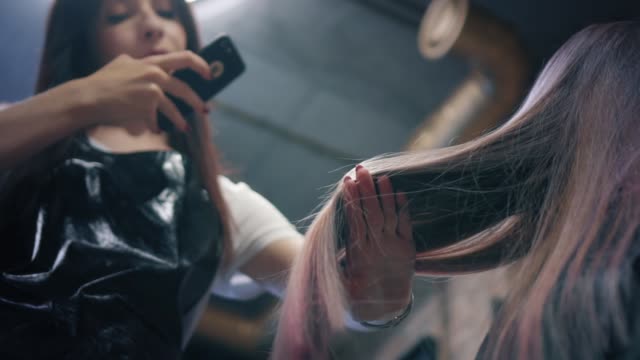 Friseur-mache-ein-Foto-von-Frau-lange-coloriertes-Haar-auf-Smartphone-im-Friseursalon.-Friseur-bewundert-ihre-Arbeit-und-zeigt-gefärbte-Haarsträhnen.-Nach-unten-Winkel.-Langsame-mo