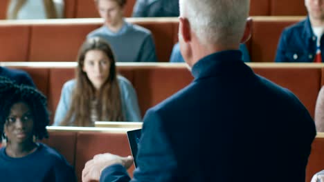 Professor-of-Computer-Science-Reads-Lecture-to-a-Classroom-Full-of-Multi-Ethnic-Students.-Teacher-Holds-Laptop-with-Deep-Learning,-Artificial-Intelligence-Infographics-on-the-Screen.