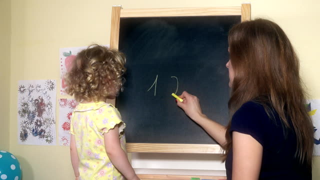 Teacher-woman-writing-numbers-on-chalk-black-board-for-little-child-girl