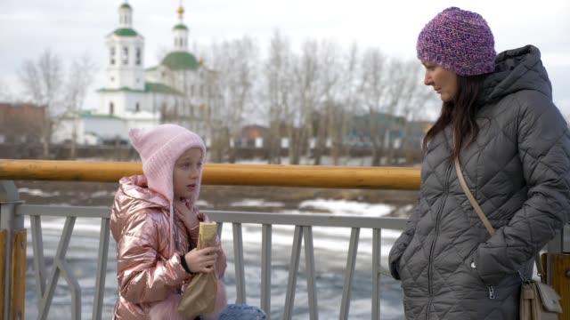 Little-daughter-eating-yummy-hot-dog-and-talking-with-mother-in-winter
