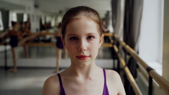 Porträt-des-jungen-Mädchens-ab-Ballett-Tänzerin-mit-Make-up-Blick-in-die-Kamera-und-lächelnd-während-Ballett-Klasse-in-geräumigen-hellen-Dancehall.-Kunst-und-Kindheit-Konzept.