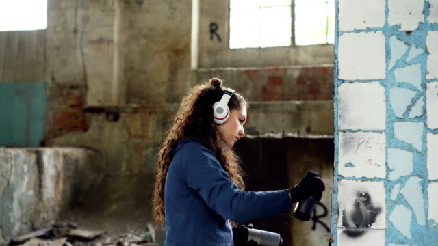 Concentrated-woman-is-decorating-old-damaged-column-inside-abandoned-industrial-building-with-graffiti-using-spray-paint-and-listening-to-music-with-headphones.