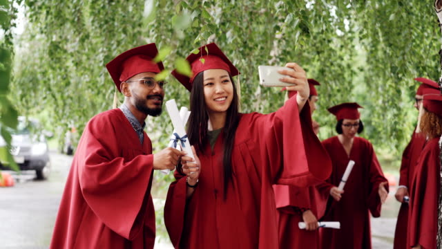 Fellow-students-are-taking-selfie-with-diplomas-posing-and-smiling,-girl-is-holding-smartphone,-people-are-wearing-gowns-and-hats.-Education-and-modern-lifestyle-concept.