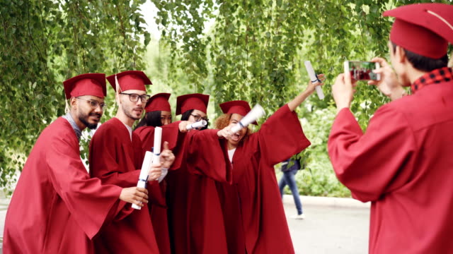 Playful-young-people-graduates-are-posing-for-photographs-making-funny-poses-and-gestures-while-their-friend-is-shooting-them-using-smartphone-camera.
