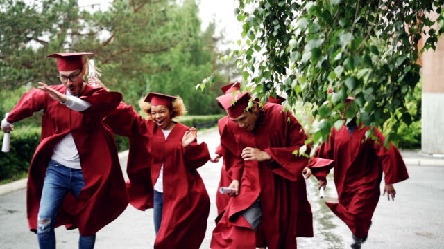 Slow-motion-of-happy-graduates-running-on-campus-waving-diplomas-and-smiling-wearing-red-gowns-and-hats.-Beautiful-trees-and-bushes-are-visible,-it-is-raining.