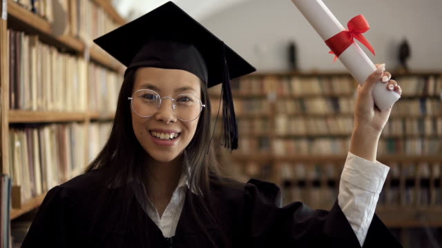 Studentin-in-akademischen-Kleid-lächelnd-posiert-positiv-stehen-in-der-Bibliothek