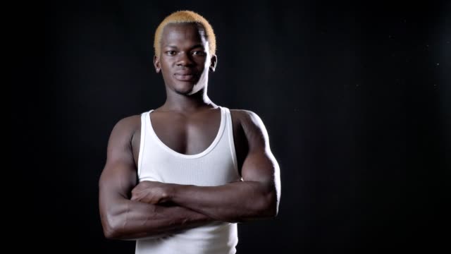 Junge-afrikanische-blonder-Mann-in-weißem-Hemd-mit-gekreuzten-Händen-stehen-und-blickte-in-die-Kamera,-selbstbewusst-und-schwere,-schwarze-Studio-Hintergrund