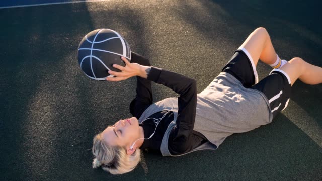 Charmante-blonde-Frau-am-Boden-liegen-und-spielen-mit-Basketball,-Musik-hören-über-Kopfhörer,-schöne-weibliche-Spieler