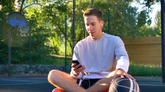 Handsome-happy-young-man-sitting-on-basketball-court-and-typing-on-phone,-holding-ball-and-listening-music,-smiling