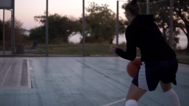 Backside-footage-of-a-young-basketball-female-player-jogging-with-ball-on-the-outdoors-court-and-throw-it-to-the-hoop
