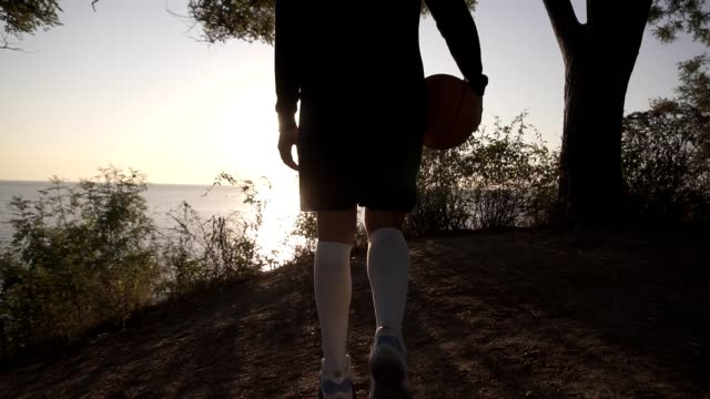 A-basketball-player-girl-comes-with-a-ball-in-her-hand,-coming-up-to-the-slope-with-trees-around.-Looks-at-the-sun-shining-over-the-sea.-Sea-view.-Backside-view