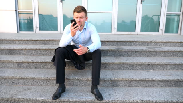 Dolly-shot-of-desperate-young-manager-talking-on-smart-phone-and-sitting-on-stairs-near-office-building.-Upset-sad-businessman-got-a-very-bad-news-on-cell-phone-and-crying-outdoor.-Close-up