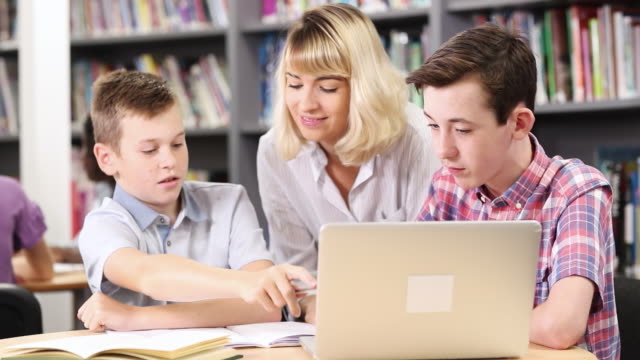 Weibliche-Lehrer-helfen-zwei-Männchen-Schülerinnen-und-Schüler-arbeiten-am-Laptop-In-der-Bibliothek