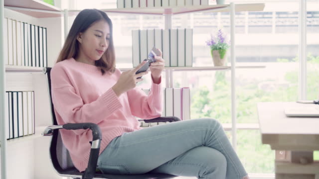 Schöne-asiatische-Frau-mit-Smartphone-kaufen-online-shopping-per-Kreditkarte-beim-Verschleiß-Pullover-sitzen-am-Schreibtisch-im-Wohnzimmer-zu-Hause.-Lifestyle-Frau-am-Wohnkonzept.
