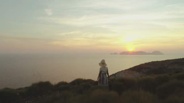 Beautiful-young-woman-with-hat-in-fashion-colorful-dress-with-skirt-and-flowers-looking-at-the-sunset-on-Ponza-Island-mountain-Italy.-Aerial-Drone-shot.