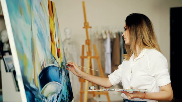 Blond-girl-painter-is-working-in-studio-depicting-sea-landscape-and-boat-on-canvas-using-brush-and-tempera-paints-looking-at-picture-and-enjoying-her-occupation.