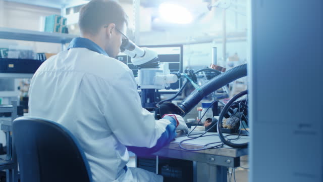 Elektronik-Fabrikarbeiter-in-weiß-Work-Mantel-inspiziert-eine-Platine-durch-ein-digitales-Mikroskop.-High-Tech-Fabrik-Anlage.