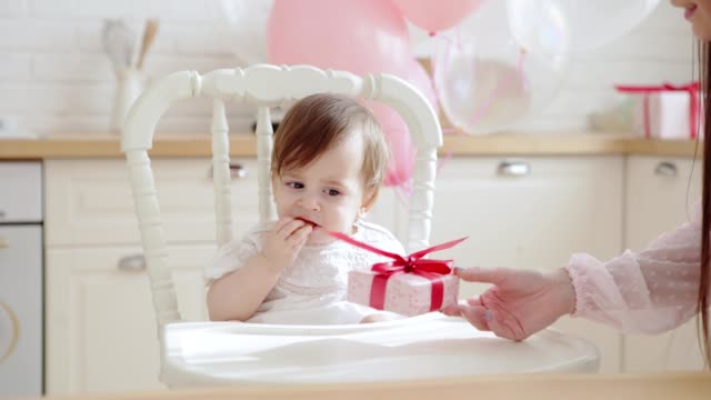 Niedliches-Mädchen-sitzt-im-Hochstuhl-und-essen-Kekse,-während-sie-Geschenkbox-am-ersten-Geburtstag-von-ihrer-nicht-erkennbaren-Mutter