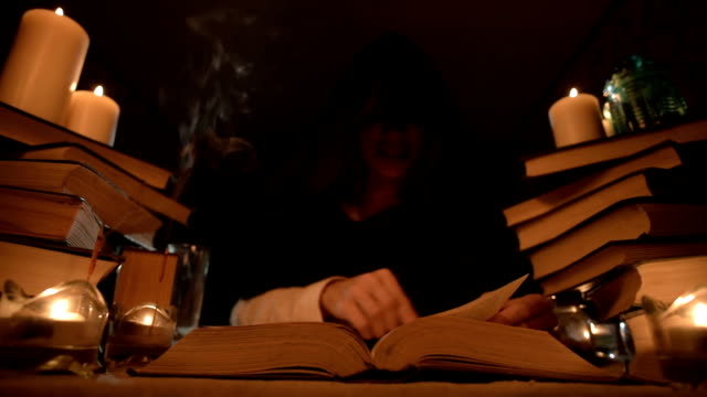 Medium-close-up-girl-magician-in-a-hood-in-a-dark-room-by-candlelight-and-looking-for-a-spell-turning-over-a-book.-Low-key.