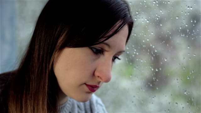 Sad-young-woman-with-absent-look-sits-by-wet-window.-Close-up