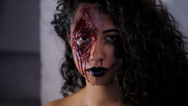 Aterrador-retrato-de-muchacha-joven-zombie-con-maquillaje-de-Halloween-sangre.-Hermosa-mujer-latina-con-cabello-rizado-en-cámara-en-estudio.-Cámara-lenta