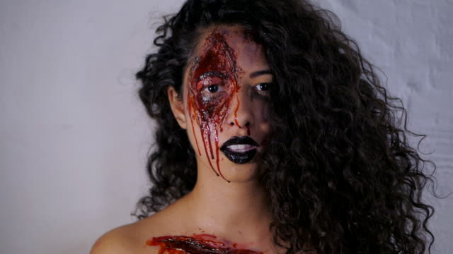 Aterrador-retrato-de-joven-asesina-con-maquillaje-de-Halloween-sangre.-Hermosa-mujer-latina-con-cabello-rizado-en-cámara-en-estudio.-Cámara-lenta