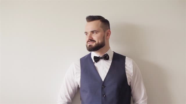 Portrait-of-30s-bearded-man-with-bowtie-stands-posing-at-white-wall-looks-at-camera-smiling.-Barber-shop-client-with-hair-gelled.-Appearance-apparel-attire-accessories-and-classic-male-fashion-details