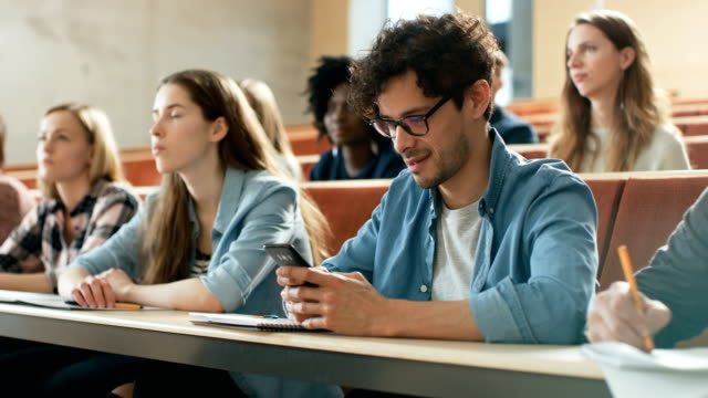 Hispanic-Mann-nutzt-Smartphone-während-einer-Vorlesung-im-Klassenzimmer.-Hörsaal-voller-Studenten.-Junge-Menschen-an-der-Universität.