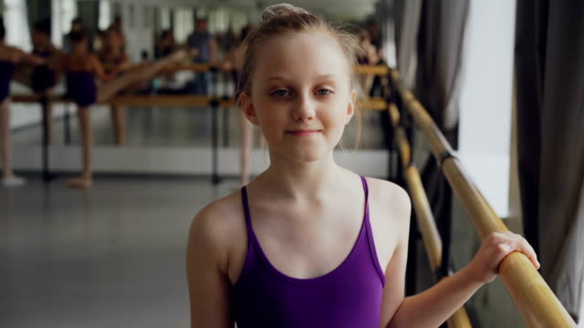 Portrait-of-beautiful-little-girl-starting-ballet-dancer-looking-at-camera-and-smiling-standing-in-ballet-class-in-spacious-light-dancehall.-Art-and-childhood-concept.