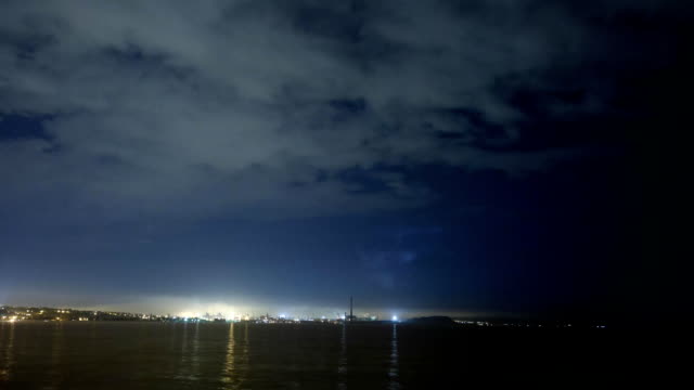 Dark-atmosphere-timelapse-of-thunderstorm-above-large-industrial-city-skyline-with-floating-clouds-and-smoke-from-pipes.-Lightning-flashes-above-the-sea-or-lake-at-night.-Ecological-problems-concept