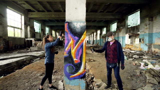 Jugendlichen-Graffiti-Künstler-sind-Aerosol-Farbe-verwenden,-um-verlassenen-Industriegebäude-mit-modernen-Graffiti-Bilder-schmücken.-Kreativität,-Straßenkunst-und-Menschen-Konzept.