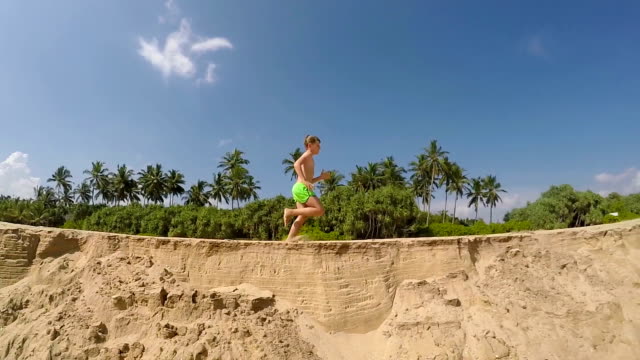 Running-little-boy-by-the-sandy-sea-beach-with-a-green-palm-trees-and-blue-sky-background-slow-motion-footage