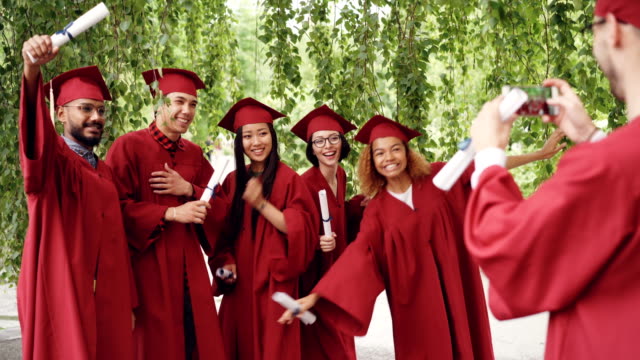 Cheerful-guy-is-using-smartphone-to-take-pictures-of-his-fellow-students-graduates-holding-diplomas,-posing-and-laughing.-Happiness,-technology-and-education-concept.
