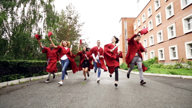 Dolly-shot-of-joyful-graduating-students-running-with-diplomas-waving-mortar-boards-and-laughing.-Higher-education,-youth-and-happiness-concept.